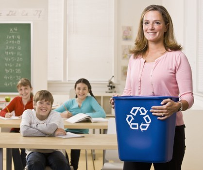 In a classroom setting, a woman stands at the front holding a blue recycling bin with a white recycling symbol, while three children sit at desks in the background, smiling and engaging with their work. The woman has shoulder-length light brown hair and is wearing a pink long-sleeved top. The children, two boys and a girl, are dressed casually and appear to be in primary school, with notebooks and pens on their desks. Behind them, a green chalkboard displays mathematical equations, and the room is well-lit with natural light coming through large windows. The environment is clean and organized, reflecting a typical school classroom. The scene subtly aligns with waste management or recycling themes, suitable for a rubbish removal service context in Wandsworth or the surrounding area.