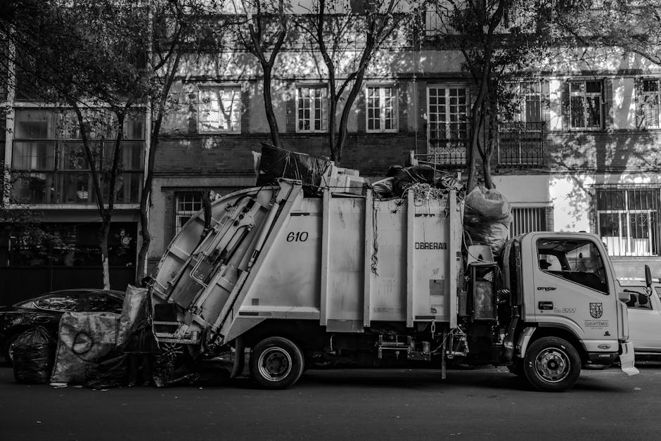 An overflowing collection of mixed waste, including paper and cardboard boxes, plastic bags, black and black-and-white garbage bags, and cardboard packaging, piled in front of a large central recyclin