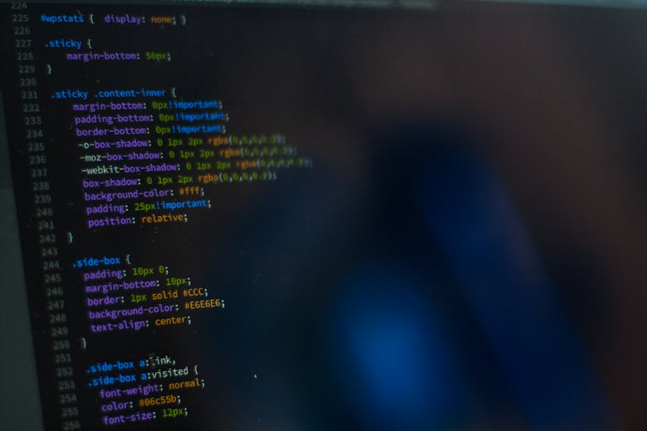 The image displays a computer screen with a dark background showing lines of colourful code and programming syntax. The code features various CSS elements, including selectors, properties, and values,