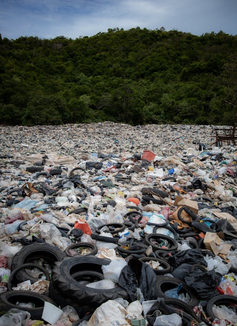 The image depicts a large, polluted landfill site with scattered waste materials covering the ground. In the foreground, numerous discarded black rubber tires are piled among crumpled plastic bags, pa